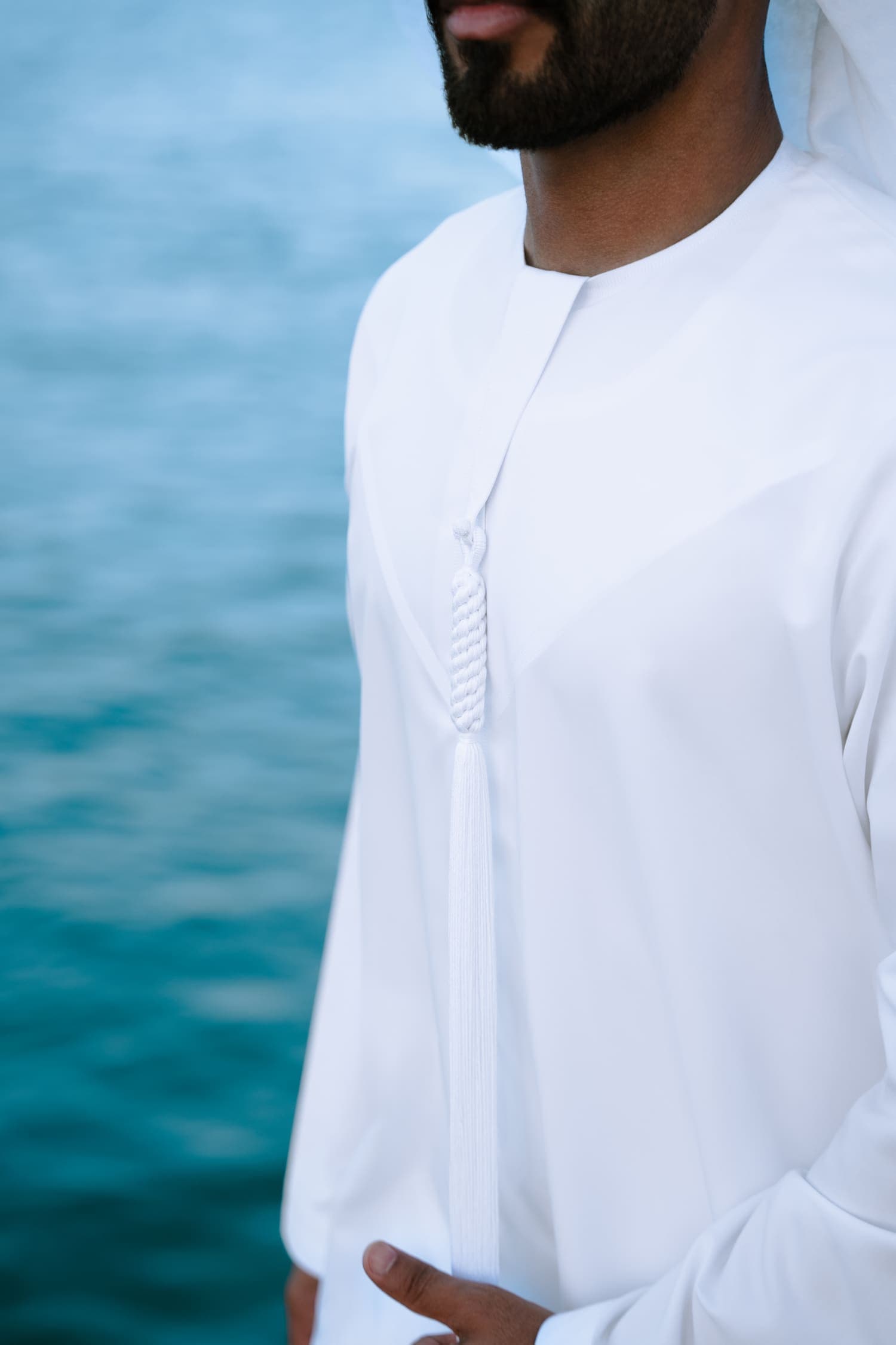 A white Misrah kandura worn by the water in Abu Dhabi, showing the collar and tarboosh tassel.